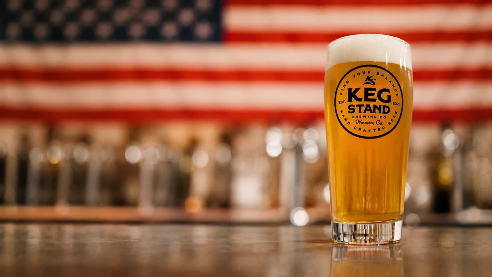 A frothy pint of Keg Stand beer in a branded pint glass on a wooden taproom bar, with a row of empty pint glasses and an American flag in the background.
